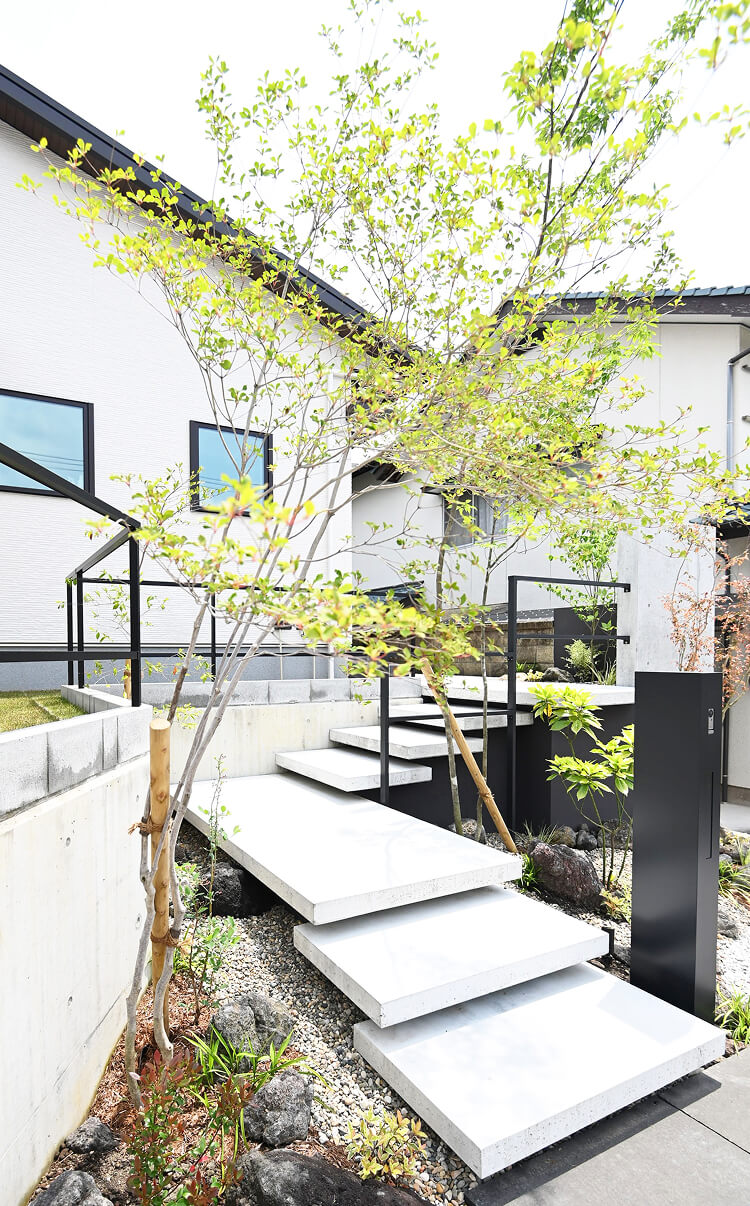 明るいアプローチに植栽を配置したモダンな外構デザイン。ステップと手すり、門柱を組み合わせたエクステリア施工例。